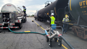 A man in safety gear climbs onto a tanker truck as a line from a tank railcar is connected; another man in safety gear watches while a third man in safety gear checks the tank railcar papers.