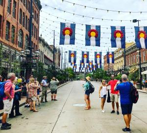A woman is talking to a group of tourists in the middle of a closed street while other tourists take photos. The Colorado State Flag flies above their heads.