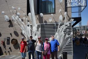 A group of people standing together getting their picture taken in front an art sculpture that appears to be milk pouring on them.