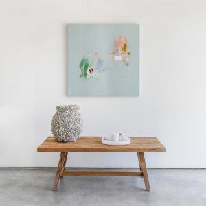 Light blue abstract painting hung about wooden table with barnacle vase sitting on top.