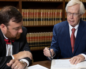 Two lawyers, wearing suits, sit at a table looking over papers with shelves of law books in the background.