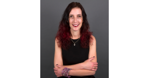 Irete Hamdani Founder of askBelynda is a woman with red hair, wise eyes and a warm smile. She is wearing glossy red lipstick and a black tank top and colourful printed pink bracelet with an unusual snake like pattern. Her nails are painted black and she w