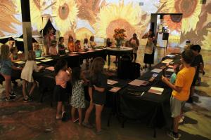 Children stand around tables drawing sunflowers