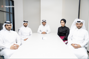 Five business people are sitting at a round conference table, smiling at the camera. This group includes four Qatari men wearing the traditional white thobe, and one Muslim woman wearing a headscarf or hijab.