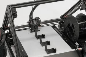 Der 3D-Fließbanddrucker von iFactory3D mit Fokus aufs Druckbett. Auf dem integrierten Förderband befinden sich mehrere technische Bauteile aus schwarzem PETG. Der Druckkopf erzeugt gerade das nächste identische Teil.
