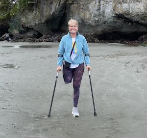 Photo of right leg above the knee amputee female on the beach wearing teal jacket and blue leggings, using forearm crutches.