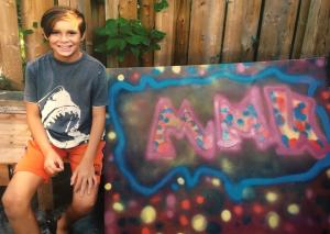 11 year old bi-racial Mexican-Canadian boy with short brown hair sitting outside beside a spray-painting canvas he created with his initials: MMK