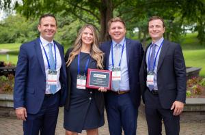 A man standing next to a woman holding a USPS Supplier Excellence Award with two other men standing on the opposite side of her. Everyone is smiling. Picture was taken in front of a lush green park with pink flowers and big trees.