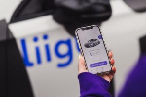 Woman is holding a phone in hand, showing the Liigu contactless car rental app, with a blurred car with Liigu logo on the background. The app is open on the pick-up screen, where the user sees their assigned car and a button "I found the car" under it. Th