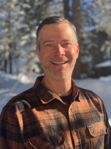 Photo of a white man with salt and pepper hair smiling at the camera, wearing an orange and black plaid shirt. Background (blurred) is the woods in winter, with snow on the trees.