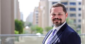 Johnny Kollin, wearing a suit, white shirt and tie, outside with buildings in the background.