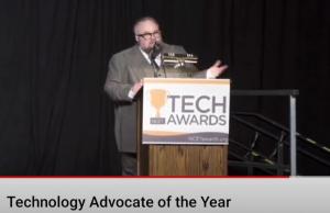 McQuillen wearing a grey suit with peak lapels speaking into a microphone, lectern has a sign that reads "TECH AWARDS" on the front
