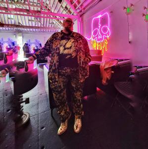 McQuillen standing in front of a pink neon skull display inside an art gallery, wearing a black and gold ornate track suit and metallic gold wingtip dress shoes