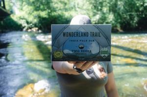 A woman standing near a river trail in Washington holding a beer box that covers her face. The beer is Wonderland Trail IPA from Two Beers Brewing and  features a design that looks like looking out from the inside of a tent onto a trail. The box features