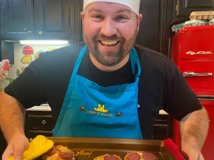 Derek Sego wears a chef hat and displays CACFP food on a baking tray.