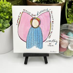 A WingTips print is displayed on a wooden easel. The whimsical angel image has playful black handwriting with splashes of color around large pink wings, “In order to realize your dreams, you must believe you are worthy of them."