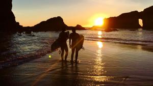 Two people with surfboards on shore with sun setting behind large rocks