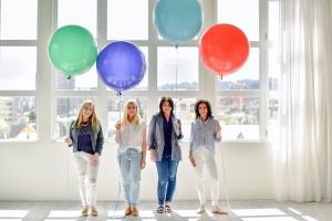 Four women stand in a line in front of a white wall with large window panes, each holding a large, colorful balloon.
