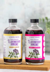 Image shows two 8 oz bottles of Carmel Berry Co's new fresh-pressed elderberry immune syrups side by side in two flavors: honey + cinnamon and honey + lemon.