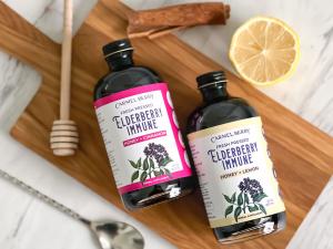 Image shows two 8 oz bottles of Carmel Berry Co's two new fresh-pressed elderberry syrups two flavors side by side: Honey + Cinnamon and Honey + Lemon.