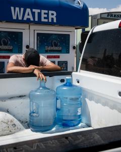 A man momentarily rests his head on his arms while filling water jugs from a paid water kiosk on a hot Texas day.