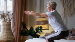 An image of an older, fit woman wearing a white t-shirt and black and white leggings with a short, blonde haircut is squatting with her arms extended in a chair exercise pose. She is in a peaceful room, with bamboo light fixtures, a palm plant in a grey p