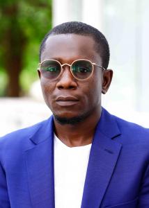 Portrait of black man with close cropped hair and lightly tinted glasses stands in the foreground with a neutral expression. He is wearing a royal blue tailored suit jacket with a white collarless shirt beneath it. The background is predominantly white an
