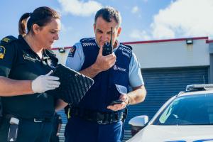 The photo shows a New Zealand ambulance officer at left speaking to a New Zealand Police officer, who is holding a Tait Communications portable radio unit.  They are shown standing alongside a Police car.