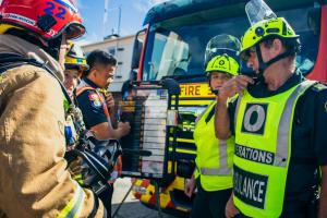 The photo shows officers from St John Ambulance and firefighters from Fire and Emergency New Zealand at a similated exercise. The group is standing in front of a fire truck and one ambulance officer is talking into a Tait Communications portable radio spe