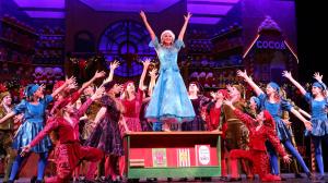 Actor is standing on a table with her arms in the air. Dancers surround her with "jazz hands". The setting is a glamorous North Pole.