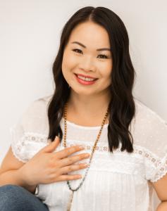 A smiling Tammy sits in a white shirt in front of a white background with her hand placed gently over her heart.