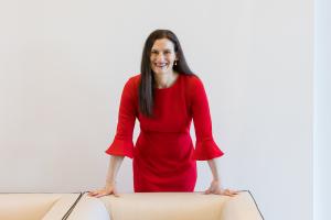 Jodi Daniels, founder of Red Clover Advisors, leans on desk wearing a red dress
