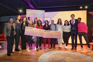 12 entrepreneurs stand on a studio stage, arms around each other, with 3 holding oversized winner's checks. They all smile excitedly.