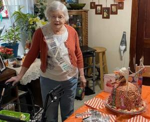 White-haird woman with one hand on her walker stands next to a table with her 100th birthday cake