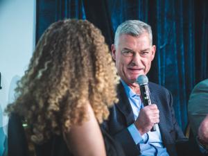 Tony Ressler is seated on a stage looking at Belva Anakwenze, the panel moderator while talking into a microphone