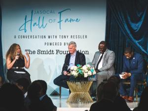 (L to R) Belva Anakwenze, Tony Ressler, Marcus Shaw, Warren Jones are seated on a stage, laughing. The stage backdrop says, JASoCal Hall of Fame, A conversation with Tony Ressler, Powered by The Smidt Foundation
