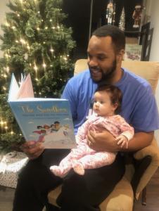 Father reading The Sandbox Series to his daughter as she sits on his lap with a Christmas tree with lights in the background.
