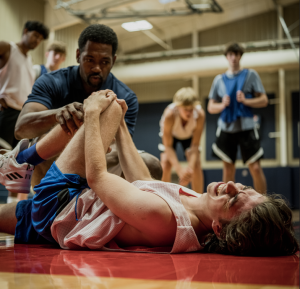 High school basketball player is injured during practice. He is lying on floor holding his knee and his coach is kneeling next to him with other players standing around looking concerned.