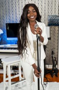 alt="Singer, Sarai Korpacz, holding mic on a stand while standing in a music studio space with piano in the background"