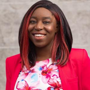 Headshot of Christine Osazuwa, a black woman with a gentle smile, a pink streak in her hair, and a bright pink blazer on