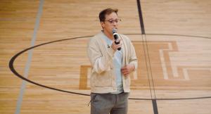 Image description for blind and low vision folks: A white man with brown hair swept one side, glasses on, and a tan colored sweatshirt cardigan stands in the middle of a school gymnasium. He is holding a microphone and speaking.