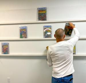 A casually dressed man facing away from the camera arranges comic books on shelves.