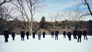 A group of 14 people, dressed in dark gray tones, stand in a brightly-lit snowy clearing surrounded by trees under a partly cloudy sky.