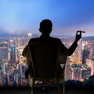 A business man sitting high above Hong Kong, smoking a cigar and looking over the city lights.