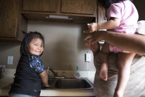 A young Navajo girl stands in front of a kitchen sink, with her hand under running water, looking back and smiling towards the camera.
