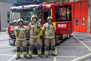 Three firefighters outside the London Fire Brigade's Dowgate station. They are standing in front of a fire truck in full operational uniform, including helmets, and their equipment includes the new radios and remote speaker microphones supplied by Tait Co