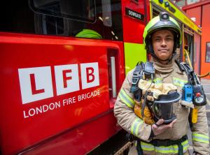 Shows a fully equipped firefighter alongside a fire truck. His equipment includes a breathing apparatus mask. The equipment includes a new radio and remote speaker microphone supplied by Tait Communications. The Tait radios are blue.