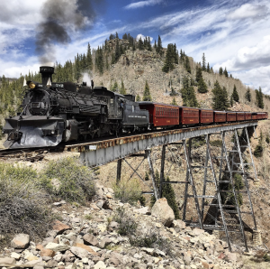 A steam engine pulling cars crosses a trestle in the mountains.