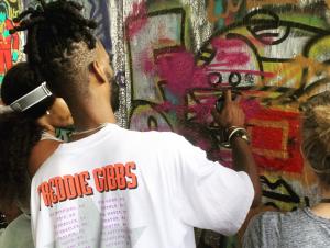 graffiti artist seen from behind sprays a wall to demonstrate to two women