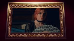A close-up of a large, horizontal shining painting hanging on a red wall. Instead of a painting, the frame holds a video still of a woman with shoulder-length ginger hair and bright red lipstick. She is holding a delicate fan and stares at the viewer accu